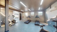 This fitness center offers a comprehensive workout facility thoughtfully organized into distinct exercise zones. The cardio area is equipped with multiple treadmills featuring integrated display consoles, a stationary bike, and a stair-stepper, all arranged on durable blue-toned flooring. Linear LED lighting runs across the ceiling, providing consistent, bright illumination throughout the space. A series of horizontal windows lines the cardio room walls, allowing natural light to filter in and offering views of the surrounding greenery.

A dedicated strength and flexibility zone features a full rack of dumbbells mirrored along one wall, stability balls, adjustable weight benches, and yoga mats laid out on the floor. A large picture window in this section frames a direct view of an outdoor swimming pool, creating an energizing and open atmosphere. Full-length mirrors amplify the sense of space and support form-checking during workouts.

An open yoga and stretching area with soft flooring provides ample room for mat-based exercises, with large windows bringing in additional natural light and outdoor views. The facility also includes a wall-mounted mini-split HVAC system for climate control, a fire extinguisher for safety compliance, and private restroom or utility access via an interior doorway. The overall design is clean, modern, and functional.