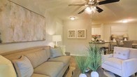 This single-floor residence offers a thoughtfully designed open-concept layout that promotes a natural flow between living and dining areas. The living room is warm and welcoming, anchored by a plush neutral-toned sofa accented with light throw pillows, and decorated with tasteful wall art that adds a relaxed, contemporary character. A ceiling fan with integrated lighting provides both comfort and ambiance, while flush-mount fixtures supplement the warm lighting throughout. The walls are finished in soft, neutral tones that complement the overall palette and create a calm, cohesive atmosphere. Decorative greenery and carefully chosen accessories bring life and texture to the space. Transitioning from the living area, an open-plan kitchen comes into view, featuring white cabinetry, dark countertops, and modern appliances, making it both functional and stylish. The kitchen bar area offers a casual seating option that connects the cooking and living spaces seamlessly. The layout is efficiently designed to maximize usability across the single floor, making the home feel spacious and easy to navigate. Overall, the residence strikes a balance between practical living and comfortable, well-curated design, making it a welcoming and functional home.