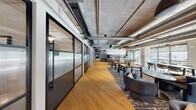 The CoWorking Area at The Smyth is a thoughtfully designed, single-floor workspace that blends industrial aesthetics with refined, hospitality-inspired interiors. Exposed concrete ceilings with visible ductwork and structural columns establish a raw, loft-like character, while warm oak hardwood flooring and carefully curated furnishings create an inviting, professional atmosphere throughout.

The open floor plan offers a variety of work settings to accommodate different needs. Communal high-top tables positioned along floor-to-ceiling windows provide ample natural light and outward views, while lounge areas furnished with leather sofas, tub chairs, and layered rugs encourage informal collaboration. A linear gas fireplace anchors one of the lounge zones, contributing to a relaxed yet productive ambiance.

Private offices and enclosed meeting rooms line one side of the floor, separated by sleek black-framed glass partition walls with frosted panels, balancing privacy with an open feel. A well-equipped kitchenette features sage green cabinetry, a quartz countertop, bold hexagonal tile backsplash, and pendant lighting. Additional breakout seating areas are distributed throughout, offering flexibility for both focused work and group gatherings. The overall design strikes a sophisticated balance between industrial structure and curated comfort.
