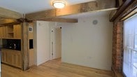 This single-floor space showcases a thoughtful blend of rustic and modern design elements. A prominent exposed wood beam runs across the ceiling, adding warmth and architectural character to the otherwise clean, contemporary interior. The ceiling is finished in crisp white, complementing the beam's natural texture and the flush-mount light fixture that provides ambient illumination.

The walls are painted a clean, bright white, creating a neutral backdrop that enhances the sense of openness throughout the space. Light-toned hardwood flooring runs throughout, contributing to the warm, inviting atmosphere while remaining practical and easy to maintain.

An in-ceiling speaker is mounted on the wall, indicating a built-in audio system for modern convenience. A doorway with a white-trimmed frame leads to an adjacent room, allowing natural light to filter through and adding depth to the layout. Additional outlets and utility connections are positioned along the walls, offering practical functionality.

The overall ambiance balances rustic charm with modern sensibility—the raw, textured wood beam contrasting elegantly with the polished floors and smooth white walls, resulting in a space that feels both timeless and contemporary.