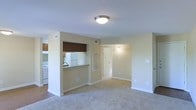This single-floor residence features an open and functional layout designed to maximize usable living space. The living area is finished with wall-to-wall carpeting in a neutral tone, creating a clean and cohesive aesthetic that complements the soft beige walls and white trim throughout. A flush-mount ceiling light fixture provides overhead illumination to the main living space.

The kitchen is positioned adjacent to the living area and is accessible via a pass-through counter with a stone-look surface, allowing for easy interaction between the two spaces. The kitchen is equipped with wooden cabinetry in a warm brown finish, a white refrigerator, and a white range. A window valance above the pass-through adds a subtle decorative touch. Hard flooring in the kitchen area contrasts with the carpeted living space.

A hallway visible from the living area leads to additional rooms, with a wall-mounted light fixture providing corridor illumination. A louvered vent panel is built into the wall along the hallway. Ample wall space and multiple electrical outlets throughout offer practical versatility for furniture arrangement and everyday use.