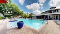 Located at 8800 Pine Forest Rd in Pensacola, Florida, this outdoor amenity space centers around a generously sized swimming pool with crystal-clear turquoise water, complemented by a separate spa. The pool deck is finished with attractive paver-style tile and is thoughtfully appointed with decorative cobalt blue ceramic planters that add vibrant color accents throughout the area. Ample lounge seating lines the perimeter, offering residents plenty of room to relax and sunbathe. Multiple shaded cabana structures with canopies, lounge chairs, and side tables provide comfortable retreats from the Florida sun. Colorful umbrellas are positioned around the deck for additional shade options. The space is framed by mature oak and palm trees that create a lush, natural canopy, lending a private and serene atmosphere. A well-maintained single-story clubhouse building with a metal standing-seam roof and large windows overlooks the pool, serving as a functional hub for the amenity area. The overall setting evokes a resort-like ambiance, combining well-designed hardscaping with verdant landscaping to create an inviting and visually appealing outdoor gathering space ideal for recreation and relaxation.