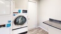 This shared laundry facility at 1840 Killingsworth Rd in Augusta, Georgia, offers a clean, functional, and well-organized space designed to accommodate multiple users efficiently. The room features light-toned walls and durable wood-look flooring that give the space a fresh, modern feel throughout.

Multiple commercial-grade Speed Queen top-load washers are arranged in a row along one wall, each equipped with card-reader payment systems for convenient, cashless operation. Alongside the washers, a bank of stacked front-load dryers lines another wall, maximizing the use of vertical space and providing substantial drying capacity. A front-load washer unit is also present, offering additional flexibility for users with varying laundry needs.

A built-in folding counter with a dark laminate surface provides a practical workspace for sorting and folding laundry, while a centrally located structural column adds architectural character without impeding the flow of the room. An informational bulletin board is mounted on the wall, keeping users informed of facility guidelines and updates.

Natural light filters in through multiple windows fitted with blinds, complemented by large flush-mounted ceiling light panels that ensure the space remains bright throughout the day. The room is accessed through a dedicated entry door, and the overall layout is open and easy to navigate, making this laundry facility a practical and pleasant amenity.