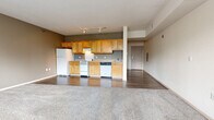 This single-floor unit offers an open-concept layout that seamlessly integrates the kitchen and living area into one cohesive space. The kitchen is outfitted with warm honey-toned wood cabinetry extending to the upper walls, providing ample storage. White appliances include a refrigerator, electric range, and dishwasher, complemented by a single-basin sink and light-toned countertops. Track lighting above the kitchen ensures well-distributed illumination throughout the cooking area, which features dark hardwood-style flooring. The living area transitions to plush wall-to-wall carpeting, creating a distinct and comfortable zone for relaxation. Neutral taupe-toned walls are consistent throughout, lending a calm and cohesive atmosphere. A pair of windows in the living space allows natural light to enter, offering views of the surrounding exterior. The textured ceiling adds a traditional character to the space. The bathroom includes a full-size soaking tub with an overhead shower, white surround, and hardwood-style flooring, finished with clean, minimal fixtures. A private entry door with an adjacent utility panel completes the functional layout of this practical and well-organized unit.