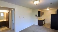 This single-floor space presents an open, functional layout that seamlessly connects the living area to the kitchen. The living space is finished with neutral wall-to-wall carpeting, contributing to a understated, practical aesthetic. White walls keep the space feeling bright and clean, while a flush-mount ceiling light fixture provides overhead illumination beneath a textured ceiling.

The kitchen area is positioned along the far wall and features dark cabinetry that offers a contrast against the light-colored countertops. A stainless steel sink is integrated into the counter, and additional upper cabinetry provides ample storage. The kitchen flooring transitions from the carpeted living area to a smooth hard surface, clearly delineating the two functional zones.

A doorway visible on the left side of the space leads to an adjacent room or hallway, suggesting additional living areas beyond what is visible. An electrical panel is mounted on the wall, and a fire extinguisher is positioned near the kitchen, highlighting practical safety considerations. The overall ambiance is clean and straightforward, offering a neutral canvas suitable for a variety of uses.