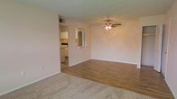 This single-floor space offers an open, functional layout that connects the living area directly to the kitchen. The living space features a combination of plush carpet and wood-look hard flooring, creating a natural transition between zones. A ceiling fan with an integrated light fixture is centrally mounted overhead, providing both ventilation and ambient lighting throughout the room. The walls are finished in a neutral, light tone that contributes to a clean and airy feel.

A pass-through opening with a small display shelf connects the living area to the kitchen, allowing for an open sightline between the two spaces. The kitchen is equipped with white cabinetry, a range with overhead microwave, and light countertops, offering a practical and well-organized cooking area. A wall-mounted HVAC vent is also visible, indicating a dedicated climate control system. The overall layout is straightforward and efficient, making good use of the available space while maintaining a simple, livable aesthetic throughout.