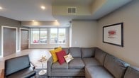 Located at 2180 Highway 13 in Mendota Heights, MN, this single-floor residence offers a thoughtfully designed open-concept layout that seamlessly connects the kitchen and living areas. The kitchen is a standout feature, showcasing warm wood-toned cabinetry paired with sleek gray countertops, a white geometric tile backsplash, and a full suite of stainless steel appliances, including a gas range, dishwasher, and French door refrigerator. Open floating shelves add both storage and visual interest against the bold dark accent wall, creating a contemporary and functional space. The living area is generously proportioned, anchored by a large sectional sofa and complemented by a glass coffee table and stylish accent chairs. Warm hardwood flooring flows throughout, tying the spaces together cohesively. Large windows allow abundant natural light to fill the room while framing views of lush greenery and a private outdoor patio or balcony beyond. Neutral wall tones with dark accent details contribute to a modern yet welcoming ambiance. Recessed and overhead lighting enhance the space throughout the day and into the evening, making this residence both practical and visually refined.