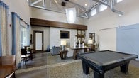This sophisticated amenity lounge is designed to impress from the moment you enter. The space is anchored by dramatic vaulted ceilings with exposed structural beams and steel accents, creating an airy, loft-inspired atmosphere. A striking stacked-stone accent wall serves as the room's focal point, framing a wall-mounted flat-screen television and providing a natural, textural contrast to the clean white walls and warm wood detailing throughout.

Natural light flows in abundantly through high clerestory windows positioned along the roofline, while a large statement pendant lantern adds a refined decorative touch overhead. The lounge area features comfortable upholstered seating arranged around a central console table, accented with table lamps, candles, and curated décor that lend a warm, welcoming ambiance.

A full-size billiards table commands the foreground of the space, making it an ideal gathering spot for social entertainment. The flooring transitions between rich dark hardwood and an ornate inlaid tile design, adding visual depth and defining distinct areas within the open-plan layout. A display shelf with framed artwork and a decorative mirror rounds out the space, contributing to its polished yet relaxed character. Overall, this lounge balances elegance with casual comfort.