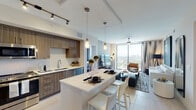 This contemporary high-rise residence offers a well-appointed, single-floor layout defined by clean lines, neutral tones, and an abundance of natural light. The open-concept kitchen is a standout feature, outfitted with rich wood-grain cabinetry, stainless steel appliances including a French door refrigerator, range, and microwave, complemented by white quartz countertops and a subway tile backsplash. A generous kitchen island with bar seating and pendant lighting serves as both a functional workspace and casual dining area, flowing seamlessly into the living room. The living area is bright and airy, anchored by floor-to-ceiling windows that frame sweeping panoramic city views. A ceiling fan, bold large-format artwork, and a mix of leather and upholstered seating lend the space a curated, contemporary feel. The primary bedroom features plush carpeting, a ceiling fan, city views, and access to a walk-in closet. A second bedroom offers tile flooring, dual round mirrors, a built-in closet, and its own access to natural light. The full bathroom includes a wood-toned vanity with a wide mirror, a combined tub and shower, and clean, neutral finishes throughout. Track lighting and pendant fixtures provide layered illumination across the residence.