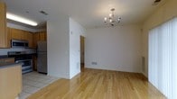 This single-story space presents a clean, open layout with a seamless flow between the main living area and the kitchen. The living and dining area is finished with warm-toned hardwood flooring that adds character and a sense of brightness to the space. A silver chandelier serves as a focal point overhead, lending a touch of elegance to the otherwise understated room. The walls are painted in a crisp white throughout, contributing to an airy and well-lit atmosphere.

A partial dividing wall separates the living area from an adjoining hallway or room, providing a degree of definition between spaces while maintaining an open feel. This passageway leads further into the unit, suggesting additional rooms beyond what is immediately visible.

The kitchen, partially visible from the main area, transitions from hardwood to tile flooring and features warm wood-toned cabinetry along with a stainless steel refrigerator. HVAC vents are integrated discreetly into the baseboards and floors, ensuring functional climate control. The overall design is straightforward and practical, with neutral finishes that offer a versatile backdrop suited to a variety of interior styles.