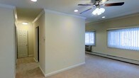 This single-floor space presents a straightforward and functional layout with a warm, neutral aesthetic throughout. The main living area is finished with wall-to-wall carpeting in a light beige tone, contributing to a cozy and comfortable atmosphere. Crown molding lines the ceiling, adding a subtle touch of architectural detail that elevates the overall feel of the space. Recessed lighting is installed in the ceiling, providing consistent ambient illumination, while a ceiling fan with an integrated light fixture offers both air circulation and additional light. A partial dividing wall separates areas of the space, creating a sense of distinct zones without fully closing them off from one another. Natural light filters in through a window dressed with sheer vertical blinds, brightening the interior during daylight hours. A baseboard heating unit runs along the exterior wall beneath the window, indicating a practical heating solution for the space. The walls are painted in a clean, off-white tone that keeps the interior feeling open and airy. Overall, the space is well-maintained and move-in ready, with functional finishes suited to everyday living.