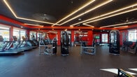 Located at 1101 S Beechwood Ave in Fayetteville, Arkansas, this single-floor fitness center is a professionally designed workout facility that combines striking aesthetics with comprehensive exercise offerings. The space is defined by a bold color scheme of vivid red walls contrasted against a dark ceiling, creating an energetic and motivating atmosphere throughout.

The ceiling is a standout architectural feature, finished in matte black and lined with geometric LED light strips that form angular, intersecting patterns, providing dramatic and even illumination across the floor. A large ceiling fan complements the lighting design while enhancing airflow throughout the space.

The fitness floor is generously sized and finished with durable rubber flooring suitable for heavy equipment use. It is divided into distinct functional zones. The cardio section lines the windowed wall and houses a row of treadmills and stair climbers, benefiting from abundant natural light streaming through large floor-to-ceiling windows that offer views of the surrounding outdoor environment. The strength training area is well-stocked with a variety of cable machines, resistance equipment, weight benches, and a dedicated free weights rack with medicine balls.

Full-length mirrors are strategically placed along the walls to enhance the sense of space and support proper form during workouts. A large flat-screen television is mounted prominently for entertainment. A separate open area near the glass entry doors provides additional open floor space, ideal for stretching or functional training. The facility presents a polished, high-energy environment designed to support a full range of fitness activities.