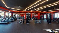 Located at 1101 S Beechwood Ave in Fayetteville, Arkansas, this single-floor fitness center is a professionally designed workout facility that combines striking aesthetics with comprehensive exercise offerings. The space is defined by a bold color scheme of vivid red walls contrasted against a dark ceiling, creating an energetic and motivating atmosphere throughout.

The ceiling is a standout architectural feature, finished in matte black and lined with geometric LED light strips that form angular, intersecting patterns, providing dramatic and even illumination across the floor. A large ceiling fan complements the lighting design while enhancing airflow throughout the space.

The fitness floor is generously sized and finished with durable rubber flooring suitable for heavy equipment use. It is divided into distinct functional zones. The cardio section lines the windowed wall and houses a row of treadmills and stair climbers, benefiting from abundant natural light streaming through large floor-to-ceiling windows that offer views of the surrounding outdoor environment. The strength training area is well-stocked with a variety of cable machines, resistance equipment, weight benches, and a dedicated free weights rack with medicine balls.

Full-length mirrors are strategically placed along the walls to enhance the sense of space and support proper form during workouts. A large flat-screen television is mounted prominently for entertainment. A separate open area near the glass entry doors provides additional open floor space, ideal for stretching or functional training. The facility presents a polished, high-energy environment designed to support a full range of fitness activities.