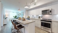 This single-floor residence is thoughtfully designed with a contemporary aesthetic, combining clean lines, neutral tones, and quality finishes throughout. The open-concept layout connects the kitchen and living area seamlessly, creating a spacious, airy atmosphere enhanced by abundant natural light filtering through large windows dressed with sheer white curtains.

The kitchen serves as a central hub, featuring glossy white and light grey flat-front cabinetry extending to the ceiling for ample storage, a classic white subway tile backsplash, and stainless steel appliances including a range, microwave, dishwasher, and refrigerator. A generously sized island with a quartz-style countertop offers seating for multiple bar stools and functions equally well for meal preparation or casual dining. Pendant lighting overhead adds a warm, modern touch.

The living area is open and inviting, with light wood-look flooring that runs consistently throughout the unit, floating shelves along the walls, and space for a dedicated workspace nook. The two bedrooms are comfortably proportioned and bright, with one featuring direct access to a private balcony with outdoor views. The full bathroom is clean and well-appointed, featuring a tub-shower combination with tiled surround, a wide vanity with under-mount sink, and matching wood-look flooring that ties the space together cohesively.