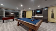 This single-floor amenity game room is thoughtfully designed to serve as a premier social and recreational destination. The space is anchored by a solid wood pool table with a bold blue felt surface, positioned at the center of the room and commanding attention as the focal point. Complementing it are a foosball table and a ping pong table, offering a well-rounded selection of classic games for guests and residents alike.

Along one wall, a built-in wet bar features dark cabinetry, a beverage refrigerator, a sink, and a wall-mounted flat-screen TV framed by decorative textured wallpaper and flanked by display cabinetry — creating an inviting entertainment hub. An accent wall in warm yellow adds a pop of color and visual energy to the room.

On the opposite end, a generous L-shaped upholstered banquette with a patterned fabric provides comfortable lounge seating, accompanied by bar-height seating and an additional wall-mounted television. The room is finished with neutral-toned carpet tiles and recessed lighting throughout, contributing to a modern, clean aesthetic.

A glass-paneled door connects the game room to an adjacent space, while a large indoor tropical plant near the entrance adds a refreshing natural element. The overall ambiance is contemporary and lively, purpose-built for leisure and social engagement.