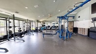 This fitness center offers a generously proportioned, single-level exercise space designed to accommodate a full spectrum of workout needs. The room features vaulted ceilings that lend an open, airy feel, complemented by recessed lighting and ceiling fans that maintain a comfortable environment during exercise. Large windows along multiple walls invite abundant natural light and provide outdoor views of the surrounding greenery, enhancing the overall atmosphere of the space.

The flooring transitions strategically between rubber sport flooring and wood-style surfaces, delineating zones for different types of training. The strength area is comprehensively outfitted with a multi-station cable machine in a striking blue finish, power racks, barbells, a full rack of dumbbells in varying weights, kettlebells, adjustable benches, and stability balls. A mirrored wall expands the sense of space and supports proper form during lifts.

The cardio zone features a lineup of StairMaster steppers, elliptical trainers, and stationary bikes, all positioned to take advantage of the natural light and outdoor views. Additional amenities include a wall-mounted television, built-in cubbies for personal storage, and a water station. The two-toned sage green and white wall treatment, combined with dark diamond-plate accent paneling, gives the facility a modern, polished aesthetic.