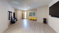 This single-level fitness studio is designed to support a wide range of exercise and movement activities. The space features light-toned hardwood-style flooring throughout, creating a clean, cohesive aesthetic that complements the neutral, warm-white walls and recessed lighting overhead.

The open floor plan provides generous room for group classes or individual workouts without feeling crowded. Along the perimeter, the studio is outfitted with a thoughtful selection of equipment, including yellow stability balls on weighted bases, a color-coded dumbbell rack, resistance bands mounted to the wall, stackable aerobic steps, and foam rollers and yoga mats stored on a dedicated rack. A wall-mounted flat-screen television is positioned for easy viewing during workouts or guided classes.

Two large botanical-themed framed artworks add visual interest to one wall, while a glass-panel exterior door allows natural light to filter into the space. A decorative multi-arm pendant light fixture provides additional ambient lighting near the entrance. The overall design is functional yet polished, with clean lines and an uncluttered layout that emphasizes versatility, making the studio well-suited for yoga, group fitness, strength training, and flexibility work alike.