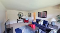 This single-floor space presents a comfortable and thoughtfully arranged living area with a clean, open layout. The living room is carpeted in a neutral tone, providing a warm foundation that complements the light-colored walls and smooth ceiling. A navy blue sofa serves as the room's centerpiece, dressed with an assortment of decorative pillows in coordinating patterns and colors. Opposite the sofa, a bold red accent chair adds a striking pop of color, positioned beside a simple floor lamp that contributes to the room's ambient lighting. A rectangular wood-and-metal coffee table anchors the seating area, styled with a red accent piece and tray for visual interest. Along one wall, a narrow console table is adorned with a sculptural circular mirror and a potted plant, adding a natural, decorative touch. Two framed artworks on the walls contribute to the space's contemporary character. A geometric blue area rug defines the seating zone, adding pattern and depth to the floor. A doorway visible in the background suggests additional rooms beyond, indicating a functional and connected floor plan throughout the space.