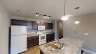 This single-floor residence in Whitsett, North Carolina offers a well-organized layout designed for comfortable everyday living. The kitchen serves as a functional centerpiece, featuring rich dark espresso cabinetry that extends from floor to ceiling, granite-style countertops, and white appliances including a range, built-in microwave, and refrigerator. A peninsula with a double-basin sink provides additional workspace and separates the kitchen from the adjoining living area. Track lighting overhead and a pendant fixture above the peninsula ensure the space is well-lit throughout. Wood-look flooring runs through the kitchen and hallway areas, adding warmth and durability underfoot.

The living area flows naturally from the kitchen and is finished with plush neutral carpet, creating a comfortable transition between spaces. Multiple bedrooms carry the same carpeted flooring and are painted in soft warm tones, each featuring windows that allow natural light to enter. One bedroom includes a ceiling fan for added comfort.

The bathrooms are finished with wood-look flooring, dark vanity cabinetry with white countertops, large mirrors, and soaking tub-shower combinations fully tiled to the ceiling. Chrome hardware and fixtures complete the clean, cohesive aesthetic throughout the unit.