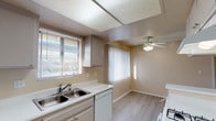 This single-floor residence offers a practical and comfortable layout with a warm, neutral color palette throughout. The kitchen is equipped with light gray cabinetry, a double-basin stainless steel sink, white countertops, and a built-in dishwasher. Natural light fills the space through horizontal blinds on the kitchen window, while a ceiling fan with a light fixture illuminates the adjacent dining area, which features light wood-look flooring. The open living area is generously sized, finished with plush gray carpet, a textured ceiling, and access to an outdoor balcony through a full-length sliding glass door with vertical blinds, offering views of the surrounding neighborhood. A wall-mounted air conditioning unit provides additional climate control. The bedroom is spacious and carpeted, benefiting from its own window for natural light. The bathroom includes a wide vanity with white cabinetry, a large mirror, and a Hollywood-style light bar, alongside a clean, white tub-shower combination and a standard toilet. A closet is accessible from the main living area, adding practical storage to the overall layout. The space presents a clean, move-in ready environment with consistent finishes throughout.