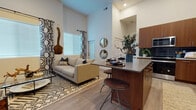 This single-floor residence showcases a thoughtfully designed open-concept layout that seamlessly connects the living, dining, and kitchen areas. Dramatically high ceilings amplify the sense of space throughout the main living area, drawing the eye upward and enhancing the airy atmosphere. Light wood-look flooring flows continuously across the space, anchoring the contemporary aesthetic.

The living area is characterized by large windows dressed with bold geometric-patterned curtains, allowing generous natural light to fill the room. A glass-top coffee table, plush sofa, and patterned area rug contribute to the modern, curated feel of the space.

The kitchen is a standout feature, equipped with rich walnut-toned cabinetry, a striking dark subway tile backsplash, stainless steel appliances including a French door refrigerator, built-in oven, microwave, and dishwasher, and a kitchen island with a gray countertop and bar seating for casual dining.

The bedroom offers a tranquil retreat with plush carpeting, a large window with blackout curtains, and ample natural light. The bathroom continues the warm wood-tone cabinetry theme, featuring a wide vanity with a white countertop, an oversized mirror, and wood-look tile flooring.