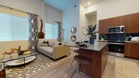 This single-floor residence showcases a thoughtfully designed open-concept layout that seamlessly connects the living, dining, and kitchen areas. Dramatically high ceilings amplify the sense of space throughout the main living area, drawing the eye upward and enhancing the airy atmosphere. Light wood-look flooring flows continuously across the space, anchoring the contemporary aesthetic.

The living area is characterized by large windows dressed with bold geometric-patterned curtains, allowing generous natural light to fill the room. A glass-top coffee table, plush sofa, and patterned area rug contribute to the modern, curated feel of the space.

The kitchen is a standout feature, equipped with rich walnut-toned cabinetry, a striking dark subway tile backsplash, stainless steel appliances including a French door refrigerator, built-in oven, microwave, and dishwasher, and a kitchen island with a gray countertop and bar seating for casual dining.

The bedroom offers a tranquil retreat with plush carpeting, a large window with blackout curtains, and ample natural light. The bathroom continues the warm wood-tone cabinetry theme, featuring a wide vanity with a white countertop, an oversized mirror, and wood-look tile flooring.