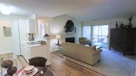 Located at 1025 Avent Hill in Raleigh, NC, this single-floor home offers a well-designed layout that balances comfort and functionality. The living area serves as the heart of the space, showcasing rich hardwood flooring that extends throughout and soaring vaulted ceilings that lend an airy, open feel. A classic fireplace with a clean white mantle serves as the focal point of the room, accented by a striking circular mirror that adds a decorative touch. Large sliding glass doors flood the space with abundant natural light and provide direct access to an outdoor patio, where views of lush surrounding greenery create a serene, private atmosphere. The open-concept design connects the living room to the dining area, which is defined by a half-wall column detail that elegantly separates it from the adjacent kitchen. The kitchen features white cabinetry and offers a pass-through view into the main living space, promoting a connected and social floor plan. Neutral wall tones throughout complement the warm wood flooring, creating a cohesive and inviting ambiance. The overall design of this home emphasizes natural light, thoughtful architectural details, and a seamless flow between indoor and outdoor living.