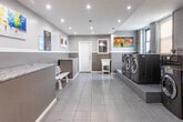 Located at 283 S Center St in Orange, NJ, this single-floor commercial laundry facility is designed for efficiency and functionality while maintaining a clean, welcoming aesthetic. The main laundry area is spacious and open, finished with large-format gray tile flooring that runs throughout the space. Multiple commercial-grade LG front-load washing machines and dryers are arranged on raised platforms, providing an organized and accessible layout for users. The exposed brick walls along one side of the room add an industrial character, complemented by painted drywall with a two-tone gray finish on the opposing walls. Recessed lighting illuminates the interior evenly, while multiple windows allow natural light to filter in. Colorful artwork displayed on the walls adds a welcoming touch to the otherwise utilitarian environment. A service counter with a dark granite top is positioned near the entrance, alongside seating for patrons. Ventilation ductwork is prominently visible near the machines, underscoring the facility's operational infrastructure. A doorway connects to an adjacent corridor, suggesting additional utility or access areas. The overall ambiance is clean, modern, and practical, making this a well-equipped and thoughtfully maintained laundry facility serving the local community.