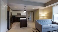 This single-floor space at Bishops showcases a contemporary open-concept design that seamlessly connects the kitchen and living area. The kitchen is well-appointed with white upper cabinetry, dark lower cabinets, and a contrasting white countertop island that serves as both a prep space and a visual centerpiece. Stainless steel appliances, including a range with a ventilation hood and a built-in microwave, add a sleek, functional touch. Pendant lighting with matte black fixtures hangs above the island, complementing the recessed ceiling lights that illuminate the space throughout. Light wood-tone flooring extends across the entire unit, enhancing the sense of openness and continuity. The living area offers a comfortable, modern feel, highlighted by warm ambient lighting from a floor lamp. A notable convenience is the in-unit washer tucked neatly into a dedicated alcove, adding practical value. The neutral wall tones — a soft gray in the main living zone and a warm olive accent — create a balanced, inviting ambiance throughout this well-designed single-level space.