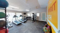 This single-floor fitness center is designed to provide a well-rounded workout environment in a compact, efficiently organized space. The room is anchored by a bold, energizing feature wall painted in vivid yellow with large motivational text reading "Energy," "Spirit," and "Strength," immediately setting an upbeat tone upon entry.

The space is equipped with multiple treadmills and a stationary bike for cardiovascular training, complemented by a multi-station cable machine and a tiered dumbbell rack for strength work. A dedicated area along one wall houses a rack of stability and exercise balls in varying sizes, alongside a water dispenser, supporting functional and flexibility training needs.

Natural light filters in through multiple windows along one wall, brightening the interior and creating an open feel. Wall-mounted mirrors expand the visual space and allow for form-checking during workouts. A mounted flat-screen television adds an entertainment option during cardio sessions. Durable, patterned carpet tiles cover the floor throughout, providing traction and comfort underfoot. Ceiling fans assist with air circulation, keeping the environment comfortable during intense activity. The overall design is practical, vibrant, and purpose-driven.