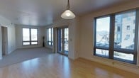 This single-floor space is characterized by its open layout and abundant natural light. The main living area features light-toned hardwood flooring that extends throughout, contrasting with a carpeted section that defines a secondary zone within the space. A pendant light fixture with a classic dome shade hangs from the smooth white ceiling, serving as a central design element.

Large windows fitted with horizontal blinds line the walls, offering views of the surrounding urban environment and distant hills or mountains. A glass-paneled door provides direct access to an outdoor balcony, further enhancing the connection to the outdoors and allowing generous daylight to flow into the interior.

The walls are finished in a neutral beige tone, contributing to a clean and understated aesthetic. White baseboards frame the perimeter of the room, adding a crisp architectural detail. The overall design is practical and uncluttered, with a layout that allows for flexible furniture arrangement. The combination of natural light, outdoor access, and varied flooring materials gives the space a functional yet inviting character suited to everyday living.