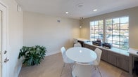 This single-floor office space is thoughtfully designed to support both collaborative and focused work. The main area centers around a round white table with modern white chairs, creating an inviting setting for meetings or group work. Along the windowed wall, a continuous built-in countertop with dark wood cabinetry below provides ample workspace and houses office equipment such as printers and a desktop computer. Large, multi-pane windows flood the area with natural light and offer views of the surrounding exterior, lending the space an open and airy atmosphere. A dedicated computer nook tucked into the corner features a built-in desk, a pendant light overhead, and a rolling office chair, offering a quieter spot for individual tasks. Light wood-toned flooring runs throughout, complementing the neutral warm-toned walls and white trim. A glass-paneled interior door connects to an adjacent tiled area, adding a sense of flow and transparency between spaces. Lush potted plants are placed throughout, introducing a touch of greenery that softens the functional aesthetic. Recessed ceiling lighting, combined with the pendant fixtures, ensures the space is well-lit at all times. Overall, the office presents a clean, modern, and practical environment well-suited for professional use.