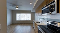 This single-floor space showcases an open-concept layout that seamlessly connects the kitchen and living area, creating a cohesive and functional environment. The kitchen is well-appointed with warm medium-tone wood cabinetry, a subway tile backsplash with subtle mixed-material detailing, and a full suite of stainless steel appliances, including an oven range, built-in microwave, and dishwasher. A double basin sink sits beneath upper cabinetry, complemented by light countertops that brighten the workspace. Light hardwood-style flooring runs throughout the space, lending warmth and continuity between zones. The living area benefits from a large window that invites generous natural light and provides views of the surrounding urban landscape. A ceiling fan with an integrated light fixture is centrally positioned, contributing to both comfort and ambient lighting. Recessed lighting in the kitchen area supplements the natural light effectively. The neutral wall tones and clean architectural lines give the space a contemporary, understated character that is both practical and inviting.