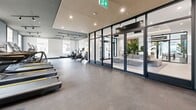 Located at 325 Royal Commons in Victoria, BC, this professionally designed fitness facility offers a generously proportioned exercise space thoughtfully laid out to accommodate a full range of workout disciplines. The interior showcases a sophisticated contemporary aesthetic, with a dark and neutral colour palette of charcoal grey tones, complemented by warm wood-slat ceiling accents and polished concrete-style flooring throughout.

The main cardio zone features a row of treadmills and stationary bikes, including spin cycles, arranged along one side of the expansive floor plate, allowing ample room for movement between equipment. A dedicated strength and free weights area houses a power rack, barbells, weight plates, dumbbells, kettlebells, and medicine balls, supported by large wall-mounted mirrors that visually expand the space and assist with form. Built-in cabinetry and open shelving provide organized storage for accessories and equipment.

Lighting is a standout feature, with track lighting running the length of the ceiling alongside recessed pot lights, creating bright, even illumination. Floor-to-ceiling glass partition walls and large windows invite natural light and maintain visual connectivity with adjacent common areas. A wall-mounted split air conditioning unit ensures year-round climate comfort. The overall ambiance is that of a boutique fitness studio — refined, functional, and inviting.