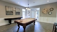 Located at 2100 Farm Springs Rd in Summerville, South Carolina, this single-floor amenity space is designed to offer residents a welcoming and functional gathering environment. The standout feature of the space is a generously sized game room centered around an ornate, dark-wood billiard table, elegantly lit by a classic pendant chandelier with frosted glass shades. Large windows throughout the room flood the space with natural light, creating a bright and inviting atmosphere. The room is finished with large-format dark tile flooring, white wainscoting along the lower walls, and neutral gray upper walls that give the space a clean, modern aesthetic. Decorative wall art and oversized clock accents add personality and warmth to the room. A secondary shuffleboard table is also visible, further enhancing the recreational appeal of the space. French doors connect the game room to adjacent areas, suggesting a thoughtful, open layout that encourages easy flow between spaces. Recessed lighting complements the natural light, ensuring the room is well-illuminated at all hours. Overall, the space strikes a balance between casual comfort and refined design, making it an ideal venue for social interaction and leisure activities.