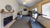 This dedicated fitness room is thoughtfully designed to support a comprehensive workout routine within a comfortable, well-appointed environment. The space is finished with warm yellow walls, wood-trimmed baseboards, and plush carpet throughout most of the floor, creating an inviting atmosphere that distinguishes it from a traditional commercial gym setting.

The room is generously proportioned, accommodating a full range of exercise equipment including a treadmill, upright stationary bike, multi-station cable weight machine, adjustable workout bench, free weights, and exercise balls. A large wall-mounted mirror reflects light and provides visual feedback during exercise. A wall-mounted flat-screen television adds entertainment during workouts, while a ceiling fan ensures consistent air circulation.

Natural light enters through double French doors that open to an outdoor patio area, as well as additional windows, keeping the space bright and airy. A fireplace with a tiled surround adds an unexpected touch of warmth and character to one end of the room. Seating, decorative plants, and accent lighting round out the space, making it as welcoming as it is functional. Baseboard heating ensures year-round comfort.