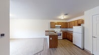 This single-floor unit offers a clean, functional layout designed for comfortable everyday living. The kitchen serves as a central hub, featuring warm honey-toned wood cabinetry extending from floor to ceiling, dark laminate countertops, a double-basin sink, a white electric range with overhead exhaust hood, and a full-size white refrigerator. A peninsula counter separates the kitchen from the adjoining living area, creating an open-concept flow ideal for both cooking and entertaining. The living space is finished with light wood-style laminate flooring that transitions seamlessly from the kitchen, while a large window at the far end draws in natural light and adds a sense of openness. The bedrooms are carpeted in neutral tones, providing a soft and quiet retreat. One bedroom features a built-in closet with a rod and shelf, while another offers access to additional storage. The bathroom is appointed with a full-size soaking tub and shower combo with a curtain rod, a vanity with a wood cabinet base, a wall-mounted mirror, and tile flooring. Throughout the unit, neutral wall colors, consistent trim work, and practical ceiling-mounted light fixtures contribute to a clean, understated aesthetic.