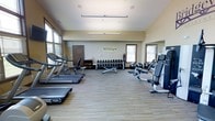 This fitness center offers a comprehensive workout environment designed to support a wide range of exercise routines. The space is outfitted with Technogym-branded equipment, reflecting a commitment to quality and performance. Along one side of the room, a row of treadmills provides ample cardio capacity, while a cable machine and adjustable bench on the opposite side cater to strength and resistance training needs. A dedicated free weights area features a full rack of dumbbells in graduated sizes, accompanied by a flat utility bench, allowing for versatile strength training exercises. Stability and medicine balls are also available, rounding out the functional fitness offerings. The room is finished with durable, wood-tone flooring that is both practical and visually clean. Natural light filters in through multiple windows fitted with blinds, creating a bright and inviting atmosphere. The vaulted ceiling adds a sense of openness and airiness to the space. Neutral wall tones and a large mirror contribute to the spacious feel of the room. A private door provides access to an additional area, and ceiling-mounted lighting ensures consistent illumination throughout. Overall, the fitness center is thoughtfully arranged to maximize usability and comfort for its users.