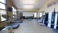 This fitness center offers a comprehensive workout environment designed to support a wide range of exercise routines. The space is outfitted with Technogym-branded equipment, reflecting a commitment to quality and performance. Along one side of the room, a row of treadmills provides ample cardio capacity, while a cable machine and adjustable bench on the opposite side cater to strength and resistance training needs. A dedicated free weights area features a full rack of dumbbells in graduated sizes, accompanied by a flat utility bench, allowing for versatile strength training exercises. Stability and medicine balls are also available, rounding out the functional fitness offerings. The room is finished with durable, wood-tone flooring that is both practical and visually clean. Natural light filters in through multiple windows fitted with blinds, creating a bright and inviting atmosphere. The vaulted ceiling adds a sense of openness and airiness to the space. Neutral wall tones and a large mirror contribute to the spacious feel of the room. A private door provides access to an additional area, and ceiling-mounted lighting ensures consistent illumination throughout. Overall, the fitness center is thoughtfully arranged to maximize usability and comfort for its users.