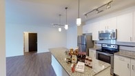 This single-floor space showcases a well-designed open-concept layout that seamlessly connects the kitchen and living areas. The kitchen is a functional centerpiece, outfitted with granite countertops that extend across both the perimeter cabinetry and a spacious island, providing ample prep and serving surfaces. White shaker-style cabinetry lines the walls, offering generous storage while contributing to a clean, bright aesthetic. Stainless steel appliances, including a refrigerator, electric range, and microwave, provide a modern, polished look. Overhead, a combination of track lighting and decorative pendant lights—suspended from the ceiling above the island—ensures thorough illumination throughout the space. The open layout allows the kitchen to flow naturally into the adjoining living area, which features warm hardwood-style flooring that contrasts attractively with the light walls and cabinetry. The neutral color palette throughout creates a cohesive, inviting ambiance. A dark-finished door visible in the background hints at additional rooms beyond, completing this thoughtfully arranged, contemporary living space.