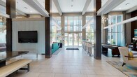 This expansive single-floor clubhouse is designed with a contemporary aesthetic that balances sophistication and comfort. The grand entrance lobby immediately commands attention with its soaring double-height ceilings, floor-to-ceiling glass walls, and large-format light tile flooring that reflects an abundance of natural light. Dark wood-clad structural columns provide a striking architectural contrast, framing views of the outdoor courtyard through the glass entry doors.

A bar-height counter with sleek white barstools runs along one side of the lobby, serving as a casual gathering spot, while a teal sectional sofa and area rug define a welcoming lounge area nearby. A shuffleboard table adds a recreational touch near the entrance.

Deeper into the space, a dedicated lounge area features rich wood-paneled walls, a large flat-screen television mounted above a warm walnut media console, and carefully curated artwork. A sectional sofa and round coffee table complete this comfortable retreat. Adjacent to this area, a modern kitchenette is outfitted with high-gloss white upper cabinetry, dark lower cabinets, a beverage refrigerator, and a white waterfall-edge island with bold yellow seating accents.

Throughout the space, recessed lighting, coffered ceiling details, and elegant chandeliers contribute to a polished, upscale ambiance suited for both social gatherings and everyday relaxation.