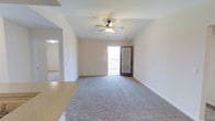 This single-floor space presents an open and functional layout centered around a spacious living area. The room is finished with neutral-toned carpet flooring that extends throughout, contributing to a clean and understated aesthetic. A ceiling fan with an integrated light fixture is centrally mounted, providing both air circulation and overhead illumination.

Natural light enters the space through a glass-panel exterior door that leads directly to a private balcony, offering outdoor access and a view beyond. The door features a sidelight panel, allowing additional daylight to filter into the interior. White painted walls and white baseboards maintain a bright, cohesive appearance throughout.

An arched passageway on one side connects the living area to an adjacent room, adding a subtle architectural detail and visual continuity between spaces. A partial view of a countertop — likely belonging to a kitchen or bar area — is visible in the foreground, suggesting a semi-open connection between the kitchen and living zones. An additional room is visible through a separate doorway in the background, indicating a straightforward, connected floor plan. The overall ambiance is neutral, clean, and move-in ready.