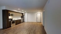 This contemporary single-floor unit offers a clean, open-concept layout that seamlessly integrates the kitchen and living area into one cohesive space. The kitchen is outfitted with a two-tone cabinetry design, pairing light upper cabinets with rich dark wood lower cabinets, complemented by stainless steel appliances including a refrigerator, dishwasher, and range. A functional sink with a sleek countertop rounds out the kitchen space. Warm hardwood-style flooring extends throughout the entire unit, adding a sense of warmth and continuity. Recessed ceiling lights provide bright, even illumination across the open floor plan, enhancing the airy atmosphere. The walls are finished in a crisp, neutral white, creating a versatile backdrop suited to a variety of interior styles. A boldly framed front entry door with a modern keypad lock system serves as a practical and stylish focal point. An adjacent hallway leads to additional areas of the unit, offering a logical and efficient flow through the space. Overall, the unit presents a modern, minimalist aesthetic with quality finishes throughout.
