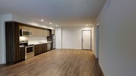 This contemporary single-floor unit offers a clean, open-concept layout that seamlessly integrates the kitchen and living area into one cohesive space. The kitchen is outfitted with a two-tone cabinetry design, pairing light upper cabinets with rich dark wood lower cabinets, complemented by stainless steel appliances including a refrigerator, dishwasher, and range. A functional sink with a sleek countertop rounds out the kitchen space. Warm hardwood-style flooring extends throughout the entire unit, adding a sense of warmth and continuity. Recessed ceiling lights provide bright, even illumination across the open floor plan, enhancing the airy atmosphere. The walls are finished in a crisp, neutral white, creating a versatile backdrop suited to a variety of interior styles. A boldly framed front entry door with a modern keypad lock system serves as a practical and stylish focal point. An adjacent hallway leads to additional areas of the unit, offering a logical and efficient flow through the space. Overall, the unit presents a modern, minimalist aesthetic with quality finishes throughout.