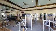 This single-floor fitness center is thoughtfully designed to support a full range of workout routines in a polished, welcoming environment. The main exercise area features a raised tray ceiling with recessed lighting, exposed dark wood ceiling beams, and a ceiling fan, creating an airy yet refined atmosphere. The space is outfitted with a comprehensive selection of Precor strength machines, free-weight stations, and a dedicated dumbbell rack, all set on durable carpet flooring. Floor-to-ceiling mirrors along the walls amplify the sense of space and allow for proper form checks during training.

A separate spin studio provides a focused environment for cycling workouts, housing multiple stationary bikes alongside a compact dumbbell rack. Natural light streams in through large windows with blinds, and the hardwood-style flooring adds a clean, modern touch to this dedicated space.

An adjacent open movement studio offers a bright, versatile area with hardwood-style flooring, large windows overlooking outdoor greenery, and a wall-mounted display screen for guided workouts. The studio's understated design keeps the focus on movement and flexibility training.

Large glass doors connect the main gym floor to the outdoors, enhancing ventilation and providing views of the surrounding landscape. The combination of high-end equipment, thoughtful zoning, and upscale finishes makes this fitness center a standout amenity.