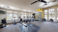 This fitness center in Humble, TX offers a well-equipped, single-floor workout space designed to energize and motivate. Entry is made through bold double glass doors etched with motivational typography, setting an encouraging tone before stepping inside. The interior is defined by an open layout with plush carpet flooring, interspersed with rubber mat sections to accommodate free weights and functional training areas.

A standout feature is the large-scale graffiti mural adorning the brick-textured accent wall, rendered in black, white, and yellow with motivational text—adding a dynamic, urban aesthetic to the space. A coordinating bold yellow accent wall further amplifies the energetic atmosphere.

The cardio zone houses multiple treadmills, ellipticals, and stationary bikes, all positioned near large multi-pane windows that flood the space with natural light and offer views of the surrounding greenery. A wall-mounted television accompanies the cardio area for entertainment during workouts. The strength training section features Hoist cable machines, a weight bench, and free weight equipment, supported by a full-length mirror wall that enhances the sense of spaciousness. Overhead, a mix of ceiling fans and contemporary multi-arm light fixtures provide both comfort and illumination throughout the facility.