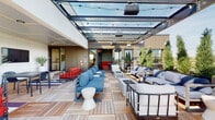This rooftop amenity level is designed to provide a dynamic combination of indoor and outdoor social spaces. The centerpiece is a spacious open-air terrace covered by a striking glass and steel canopy, which allows natural light to flood the space while providing shelter from the elements. Strung with café lights and equipped with overhead heaters, the terrace creates an inviting atmosphere day or night. The teak-style deck flooring ties the space together, hosting multiple distinct seating areas furnished with blue and neutral-toned sectional sofas, fire pit tables, and modern accent stools.

The terrace transitions to a semi-covered area featuring bold red outdoor sofas, wall-mounted televisions, and tall potted plants, creating a vibrant lounge environment ideal for gatherings. A dedicated interior lounge area connects seamlessly to the outdoor space and is distinguished by eye-catching teal geometric-patterned wallpaper, warm wood-slat ceilings, and woven pendant light fixtures. Red leather banquette seating paired with marble-top tables and sculpted barrel chairs adds a retro-modern flair. A small kitchenette with dark cabinetry, a marble countertop, and gold fixtures completes the interior. The entry lobby features large-format tile flooring, vertical wood paneling with integrated lighting, and textured wallcoverings, establishing a polished and cohesive design identity throughout.
