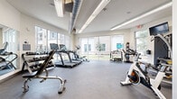 This modern fitness center is designed to support a full range of workout routines in a bright, open environment. The space is defined by large floor-to-ceiling windows that line multiple walls, flooding the interior with natural light and offering views of the surrounding outdoor grounds. The durable rubber flooring throughout provides a safe, functional surface for all types of exercise.

The cardio zone features multiple treadmills equipped with individual display screens, as well as stationary and spin bikes, offering members a variety of aerobic training options. A well-organized free weight station includes a tiered dumbbell rack and adjustable benches, complemented by a large wall mirror that enhances the sense of space and allows users to monitor their form. Wall-mounted televisions are positioned throughout for entertainment during workouts.

A dedicated functional training area provides open floor space alongside a medicine ball rack, resistance accessories, and additional equipment for dynamic exercise. Exposed ductwork on the ceiling adds an industrial aesthetic that contrasts cleanly with the bright white walls.

The fitness center also features a glass-walled partition offering views into an adjacent lounge or amenity space, a wall-mounted water fountain, and a hand sanitizer station. Clearly posted fitness center guidelines and signage round out this thoughtfully appointed facility.