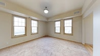This single-floor space presents a clean and well-lit interior defined by its warm, neutral aesthetic. The primary room is generously sized, featuring plush wall-to-wall carpeting in a soft, neutral tone that adds comfort underfoot and contributes to the room's inviting atmosphere. A notable tray ceiling adds architectural interest and a sense of added height to the space, while a centrally mounted ceiling light fixture provides practical overhead illumination.

Natural light is a highlight of this space, with multiple double-hung windows framed in warm wood-toned trim lining two walls. These windows allow abundant daylight to fill the room, creating a bright and airy environment. The windows also offer views to the exterior, further enhancing the open feel of the space.

The walls are finished in a soft, warm beige palette that complements the carpeting and window trim, creating a cohesive and understated interior. Visible HVAC vents are integrated into both the ceiling and walls, ensuring climate comfort throughout. The overall design is straightforward and functional, presenting a versatile, move-in-ready space well-suited for a variety of uses.