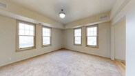 This single-floor space presents a clean and well-lit interior defined by its warm, neutral aesthetic. The primary room is generously sized, featuring plush wall-to-wall carpeting in a soft, neutral tone that adds comfort underfoot and contributes to the room's inviting atmosphere. A notable tray ceiling adds architectural interest and a sense of added height to the space, while a centrally mounted ceiling light fixture provides practical overhead illumination.

Natural light is a highlight of this space, with multiple double-hung windows framed in warm wood-toned trim lining two walls. These windows allow abundant daylight to fill the room, creating a bright and airy environment. The windows also offer views to the exterior, further enhancing the open feel of the space.

The walls are finished in a soft, warm beige palette that complements the carpeting and window trim, creating a cohesive and understated interior. Visible HVAC vents are integrated into both the ceiling and walls, ensuring climate comfort throughout. The overall design is straightforward and functional, presenting a versatile, move-in-ready space well-suited for a variety of uses.